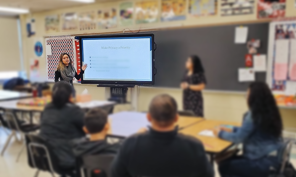 image description: classroom SMART board presentation on social media in classroom, two facilitators standing in front, with seated attendees